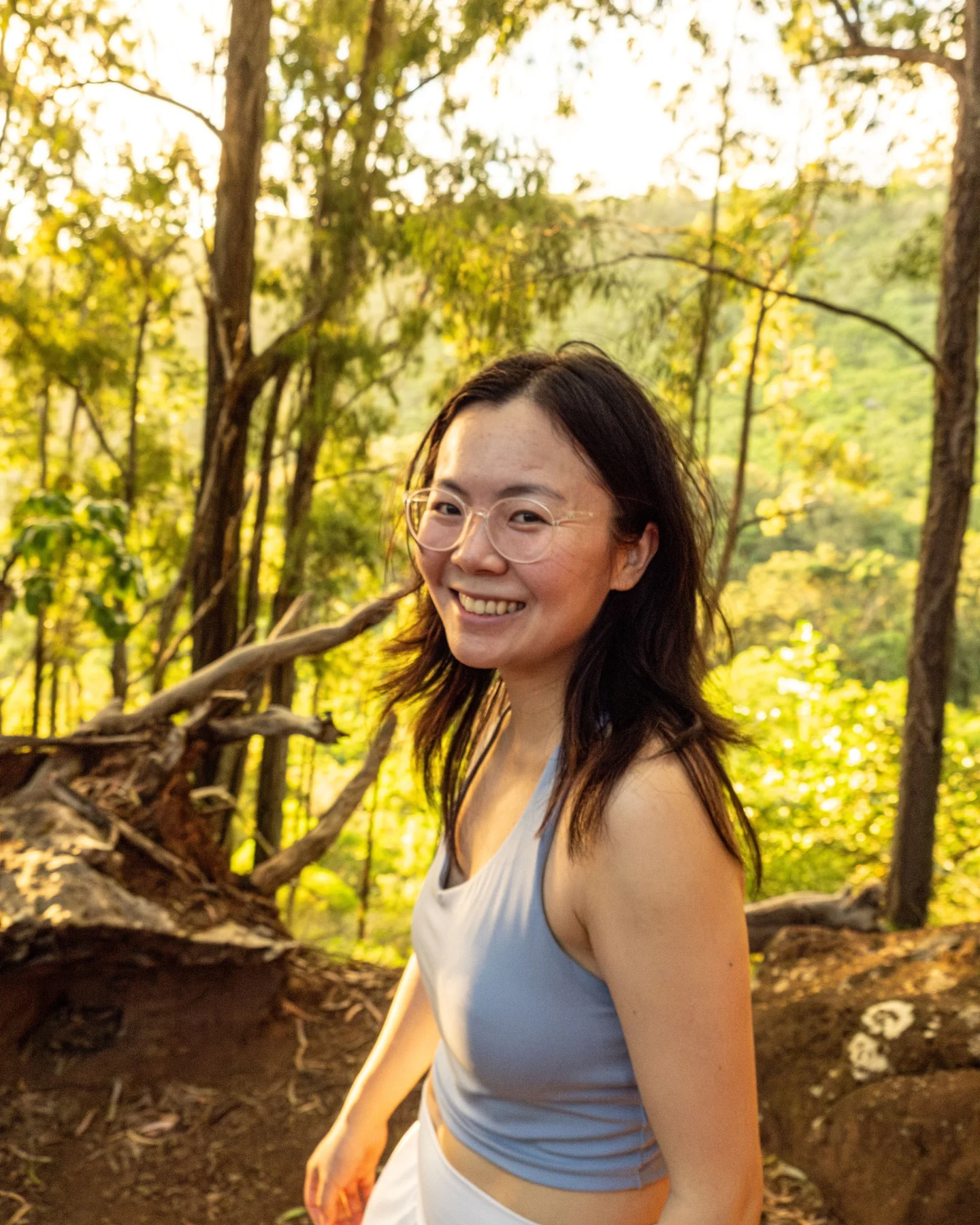 Smiling woman with glasses in a sunlit forest.