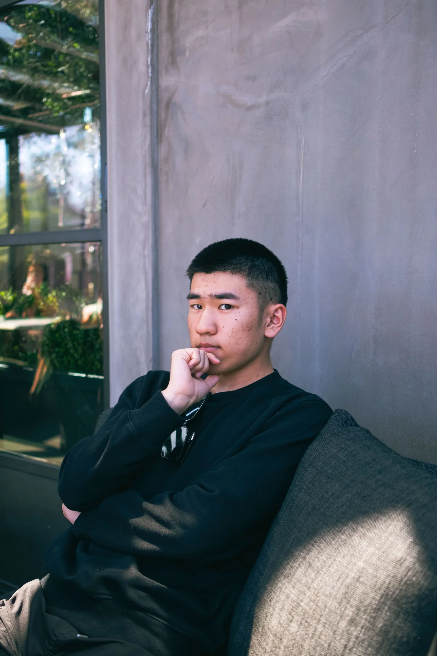 Young man in a black sweatshirt seated against a gray wall.