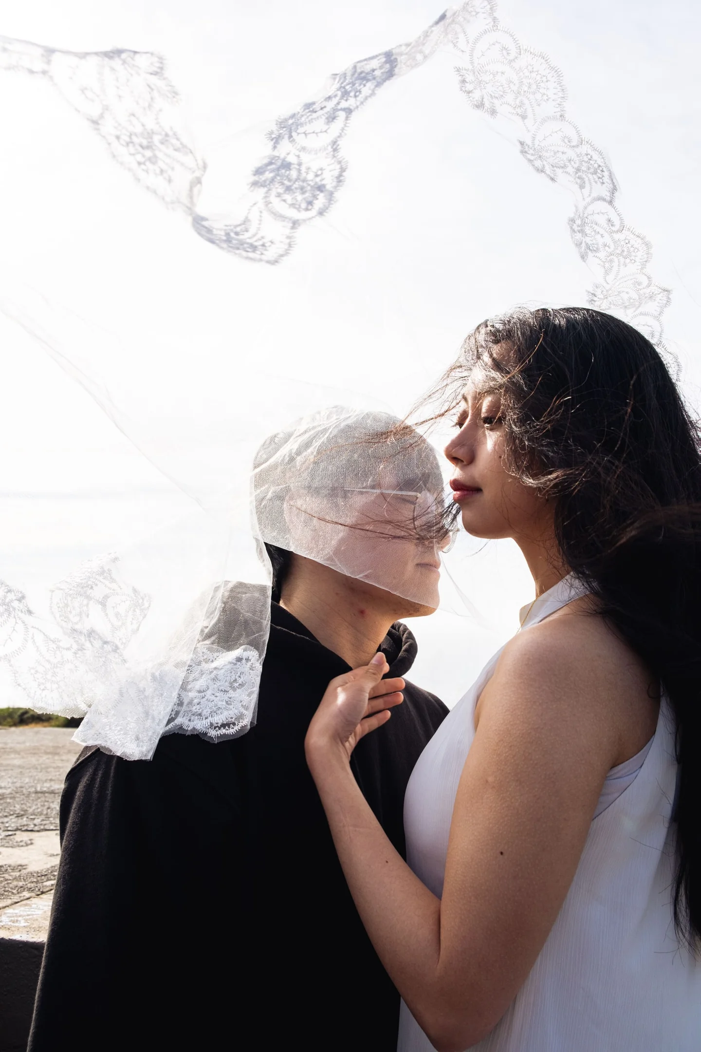 Two people standing close beneath a lace veil in the wind.
