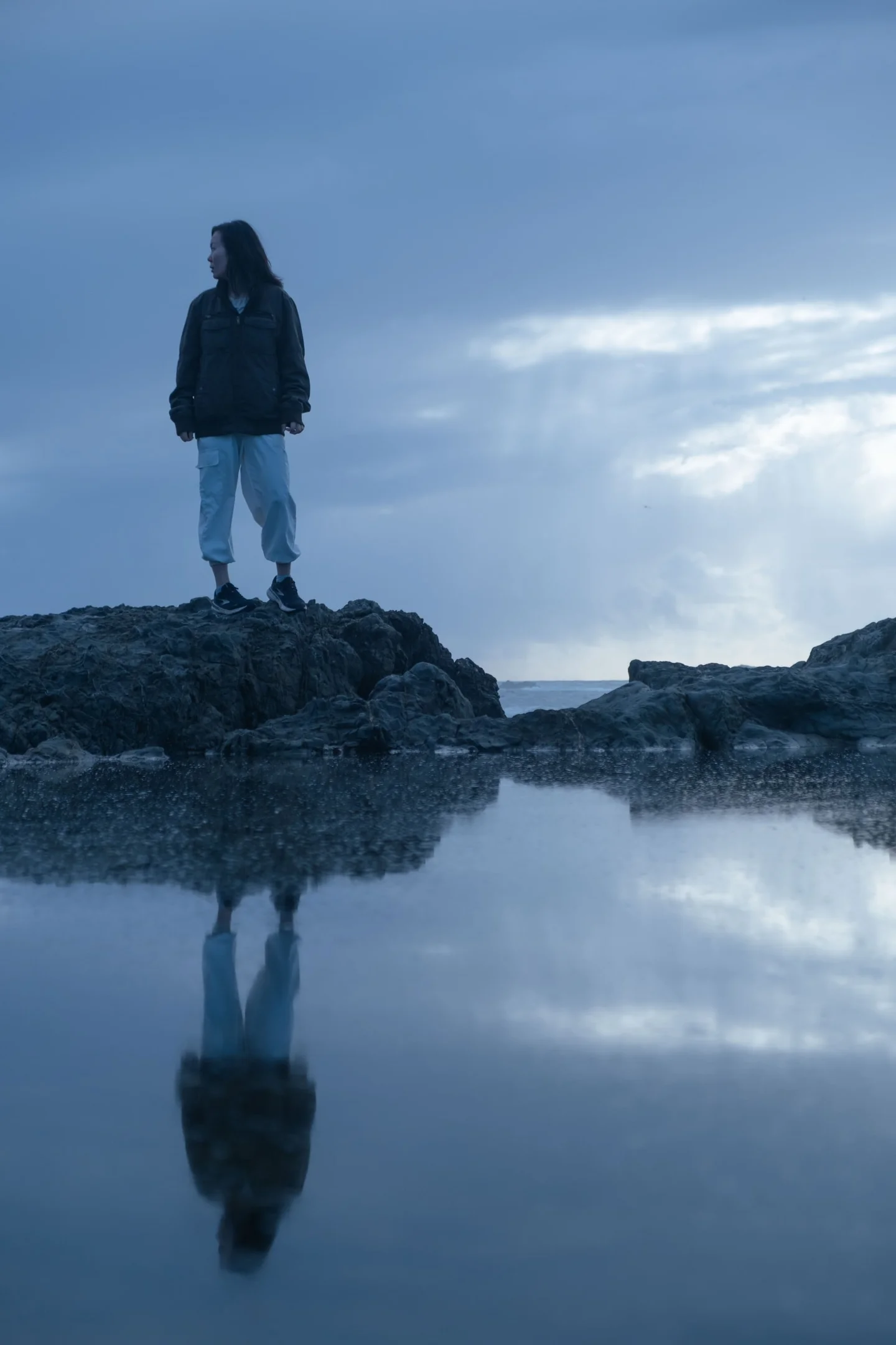 Woman standing on coastal rocks above her reflection in a tide pool.