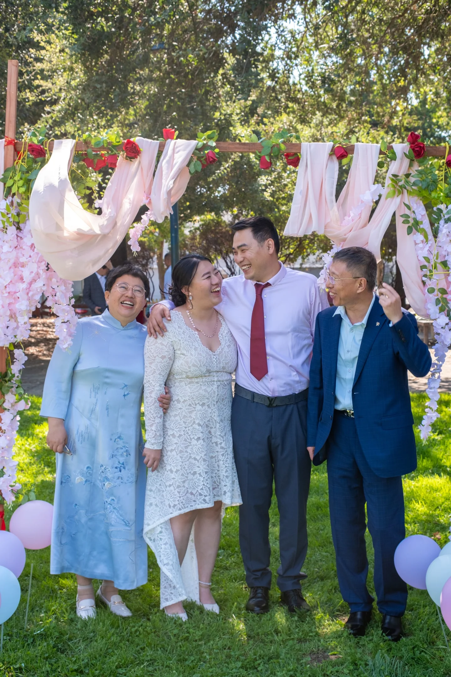 Four people laughing together beneath a flower-decorated wedding arch.