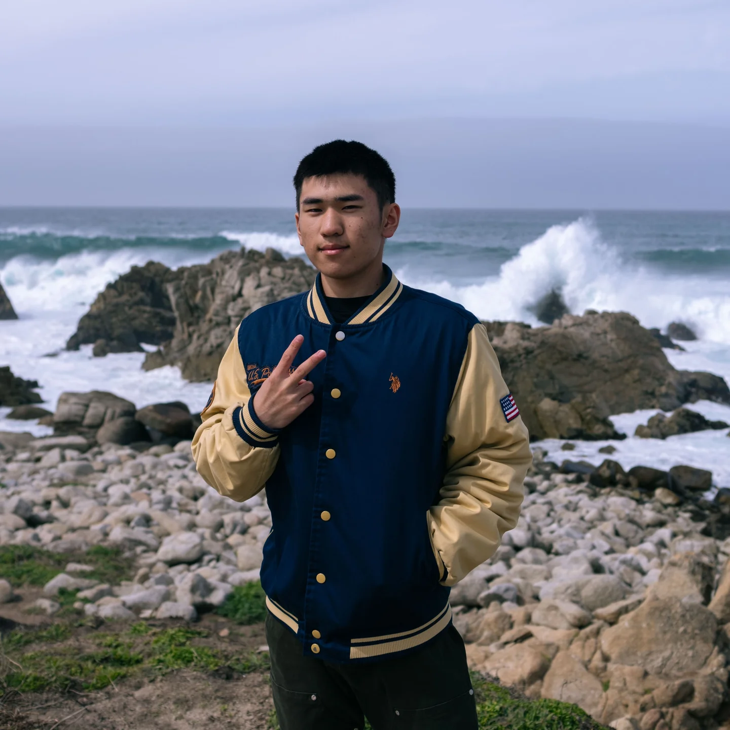 Man in a blue and gold varsity jacket beside a rocky shoreline.