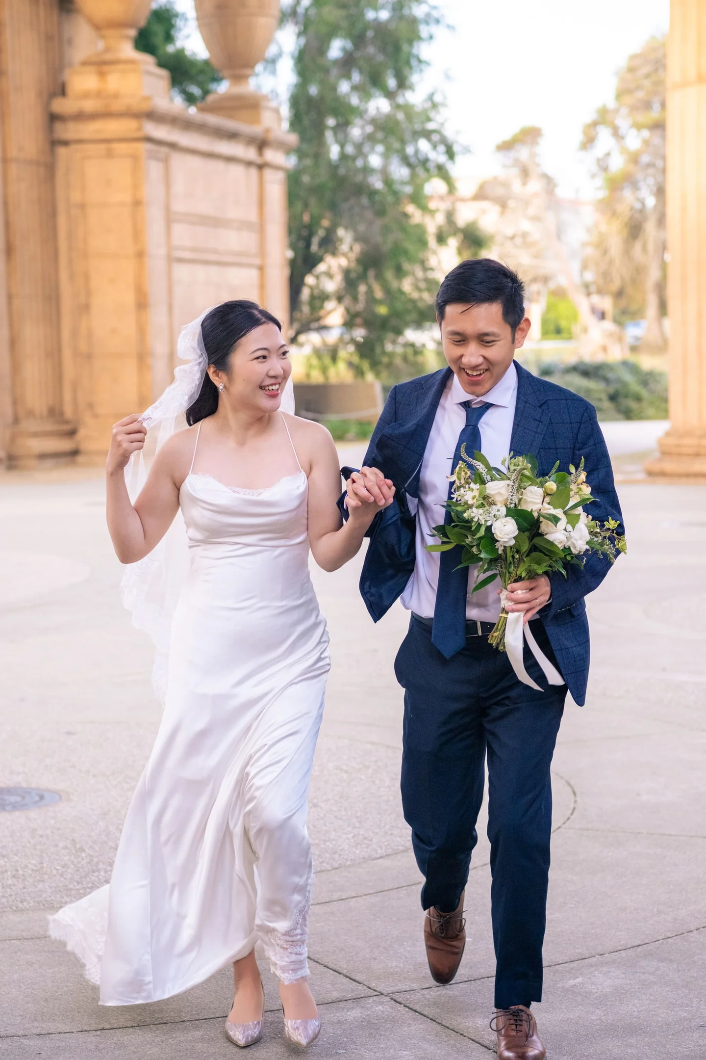 Couple smiling and running hand in hand beneath stone columns.