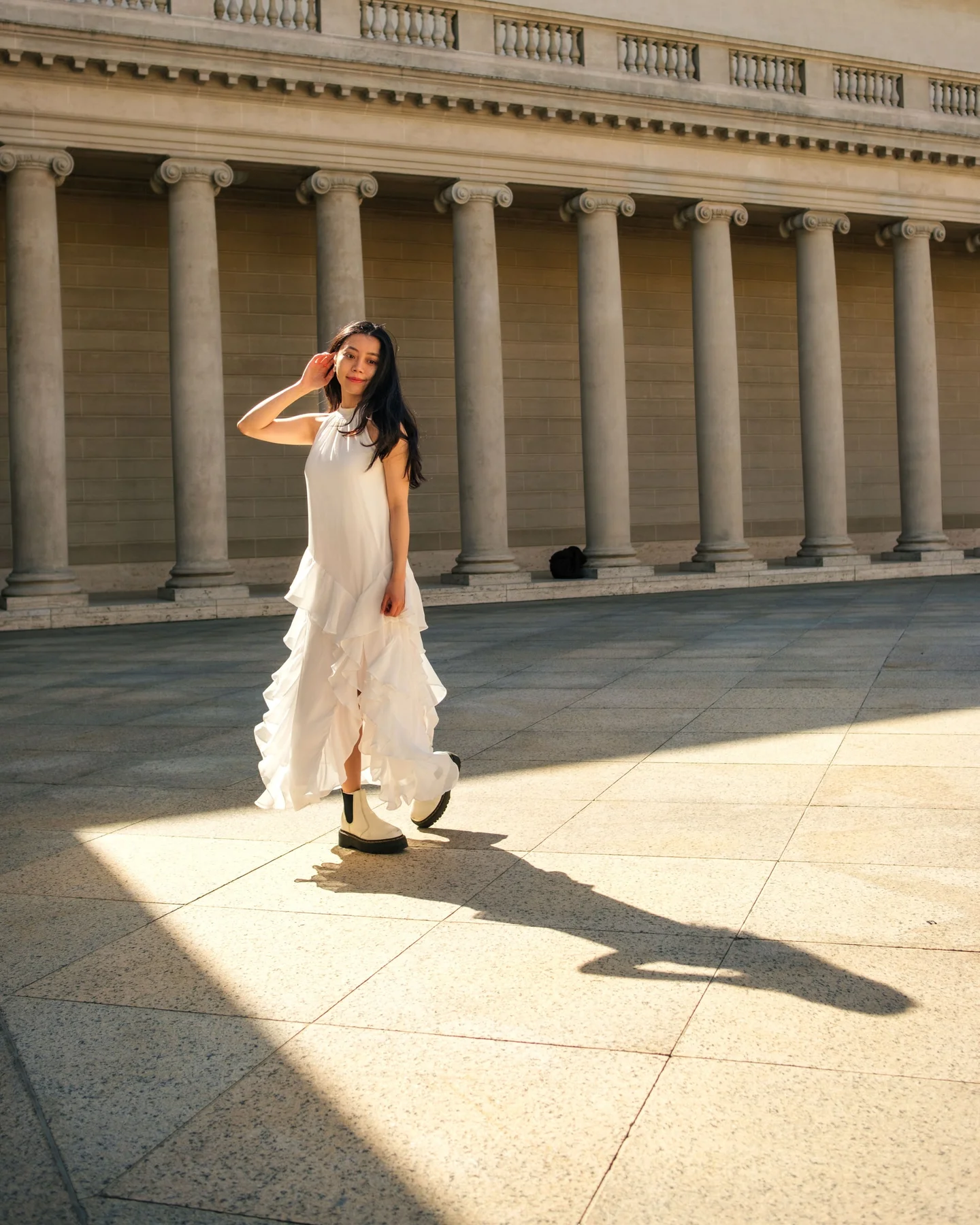Woman in a white dress standing in sunlight in front of a colonnade.
