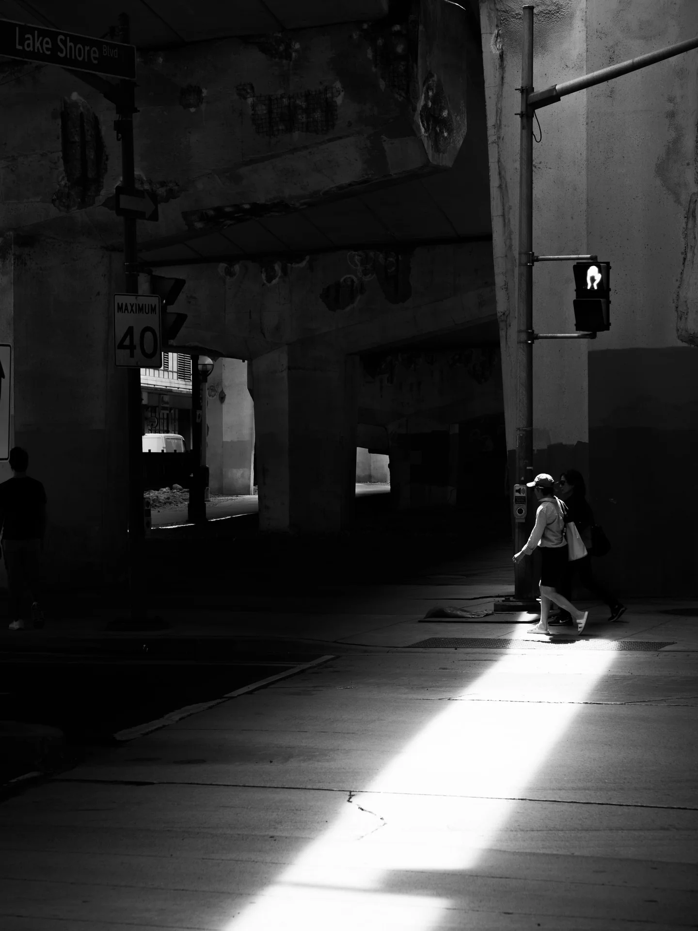 Two pedestrians crossing a bright strip of sunlight beneath a concrete overpass in black and white.