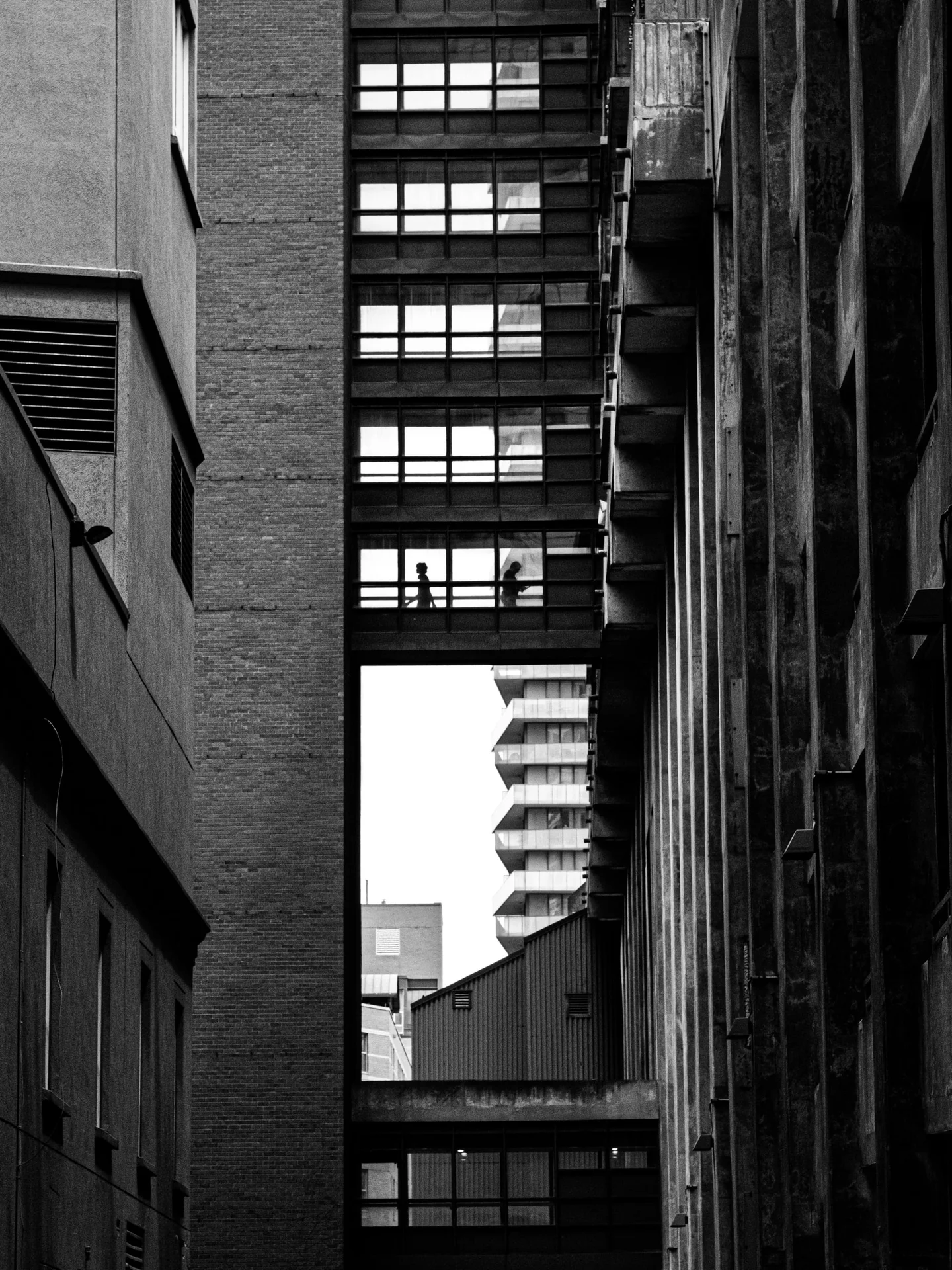 Two silhouettes inside a glass walkway between tall buildings in black and white.