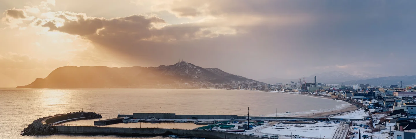 Snow-covered coastal town and harbor beneath mountains in golden evening light.