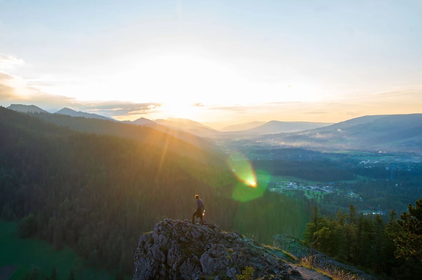 Small figure on a rocky outcrop overlooking a forested valley at sunset.