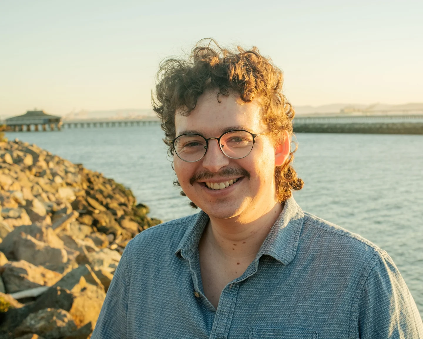 Smiling man with glasses beside the water at sunset.